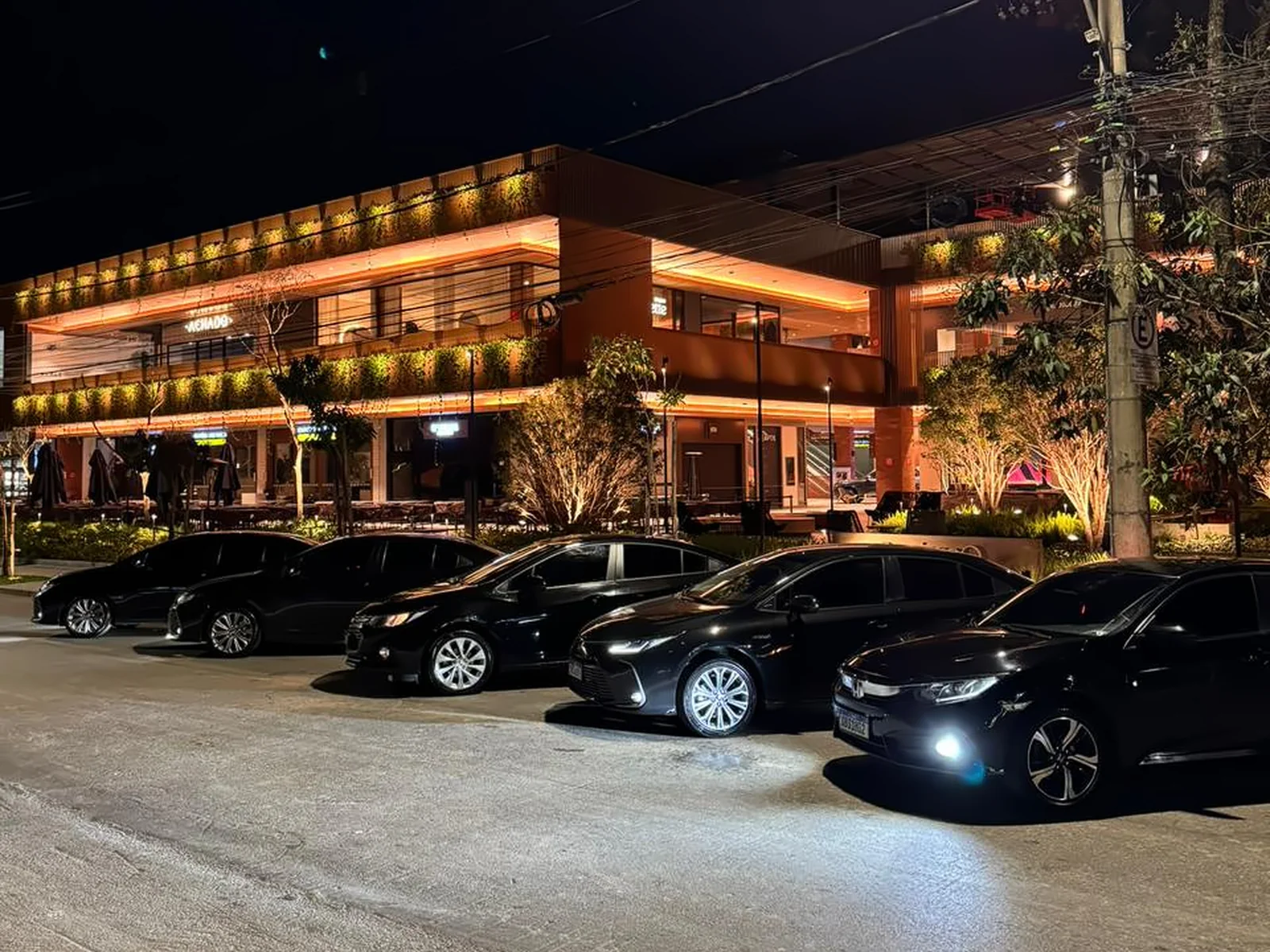 Frota de sedans executivos pretos da Elite enfileirados à noite em frente a restaurante iluminado